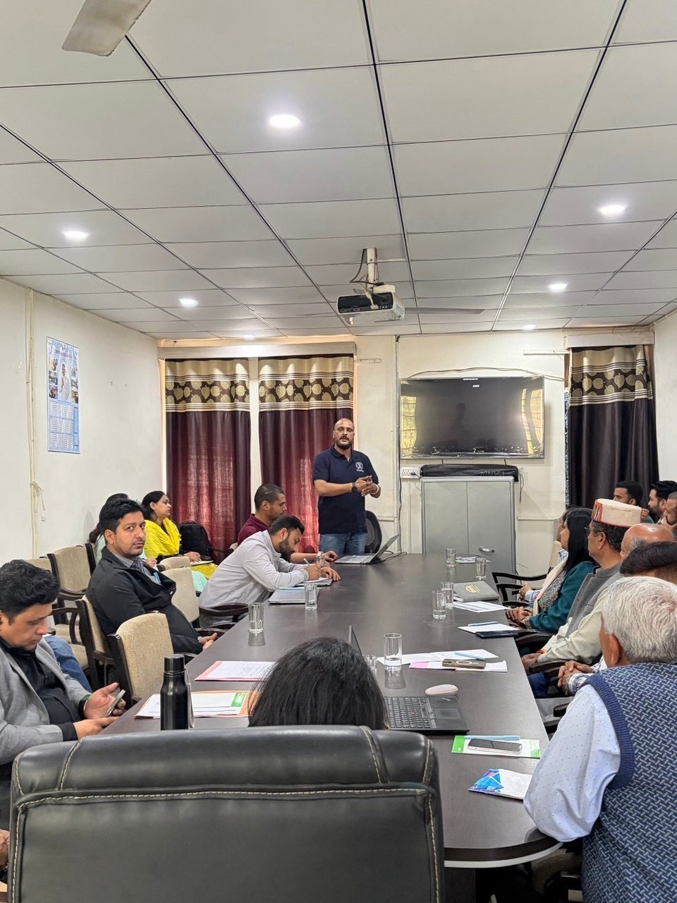 A briefing in progress at the Sub-Divisional Magistrate's office in Mandi. A member of the iIoTs team stands at the head of a long conference table addressing around a dozen seated officials, several with papers and notes in front of them and one wearing a traditional Himachali cap. A ceiling projector and a wall-mounted screen are at the far end of the room.