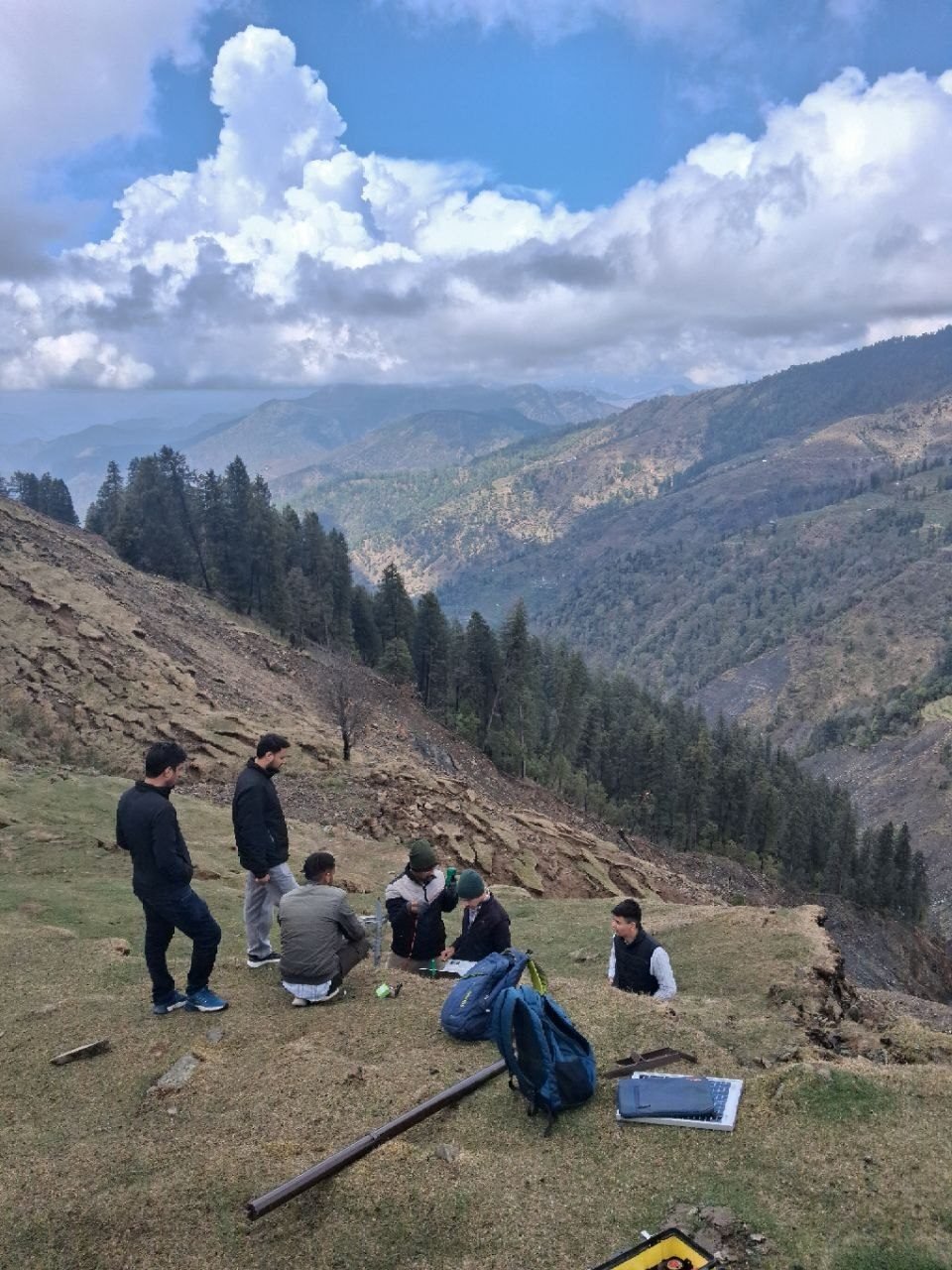 Six members of the iIoTs field team on a grassy ridge at Parashar, four of them crouched over equipment and an open case, with a steel pole lying ready on the ground, an eroded slope behind them and a forested valley beyond.