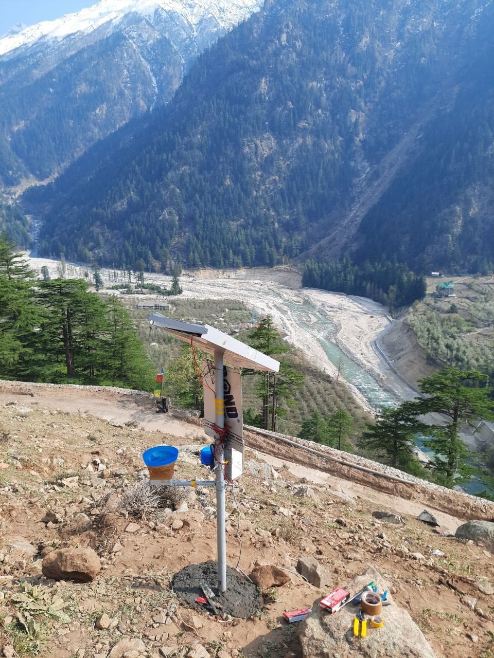 A solar-powered monitoring station on a bare ridge: a steel pole set in fresh concrete carrying a solar panel, a sensor enclosure and a blue funnel rain gauge, with a forested Himalayan valley, a wide braided riverbed and snow-covered peaks behind it.